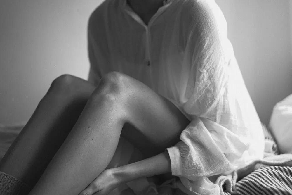 黑白照,穿寬鬆白襯衫的女性坐在床上,雙腿彎曲交疊,一手輕放在小腿上,畫面聚焦在膝蓋